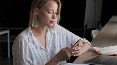 A serious focused young blonde woman typing on her smartphone while sitting near the window indoors