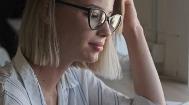A beautiful young woman wearing glasses is using her smartphone while sitting near the window indoors