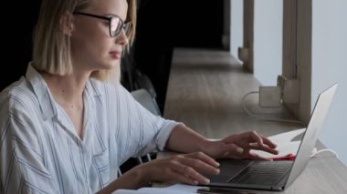 A serious calm young businesswoman wearing glasses is using her laptop while sitting near the window in the office indoors