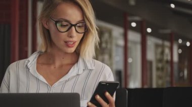 A laughing young woman wearing eyeglasses is using a laptop and cellphone while sitting in conference hall