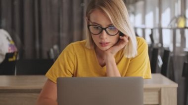A pensive calm young woman wearing eyeglasses is working on her laptop while sitting at the desk in a conference hall