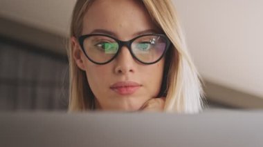 A close-up view of a nice focused woman wearing eyeglasses is working on her laptop while sitting at the desk in a conference hall