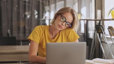 A focused blonde woman wearing eyeglasses and yellow t-shirt is working on her laptop while tilting her head in a conference hall