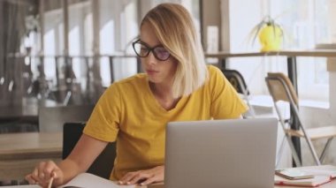 An attractive young woman is making notes on her notebook while working on her laptop in a conference hall