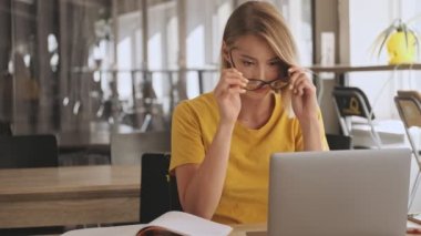 A pretty young woman is putting on her eyeglasses while making notes on her notebook and working on her laptop in a conference hall