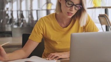 An attractive young woman wearing eyeglasses is making notes on her notebook while working on her laptop in a conference hall