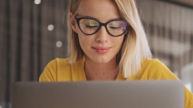 A close-up view of a smiling blonde woman wearing eyeglasses is working on her laptop in a conference hall