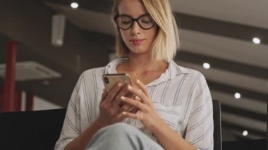 A good looking calm woman wearing eyeglasses is using a phone while waiting in a conference hall