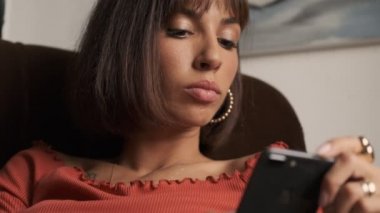 Pensive cute caucasian young woman in a brown t-shirt surfing on phone and looking around while sitting in an armchair