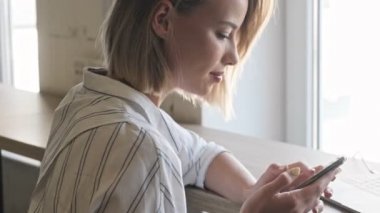 An attractive blonde woman is using the smartphone while sitting at the desk near the window