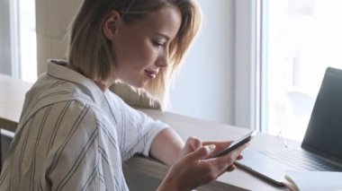 A nice blonde woman is using the smartphone while working on the laptop near the window