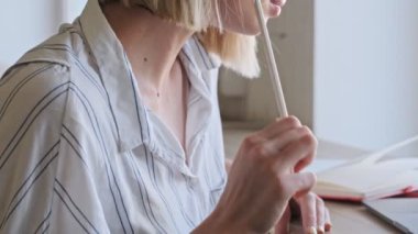 A close up view of a nice blonde woman is working with a notebook and a laptop in the office near the window