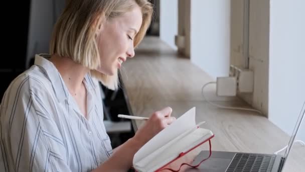 Egy Koncentrált Szőke Valamit Jegyzetfüzetébe Miközben Egy Laptopon Dolgozik Ablakhoz — Stock videók