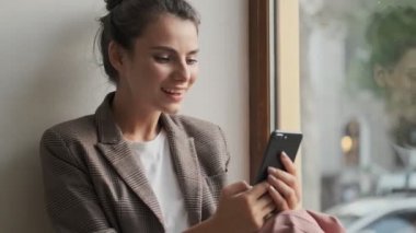 Çekici, gülümseyen genç bir kadın pencerenin yanında otururken akıllı telefonunu kullanıyor.