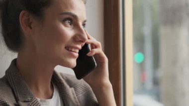 Güzel, şık bir genç bayan pencerenin yanında oturan biriyle telefon görüşmesi yapıyor.