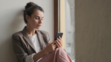 Genç bir bayan pencerenin yanında otururken akıllı telefonunu kullanıyor.