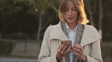 Açık hava havaalanı yakınlarında akıllı telefon kullanan ceketli, sırıtan sarışın iş kadını.