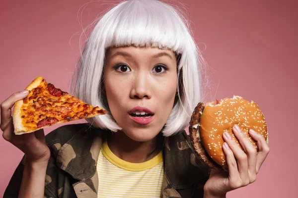 Imagen de chica asiática joven sosteniendo rebanada de pizza y hamburguesa grande —  Fotos de Stock