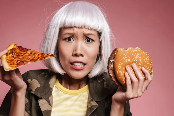 Imagen de chica asiática joven sosteniendo rebanada de pizza y hamburguesa grande — Foto de Stock
