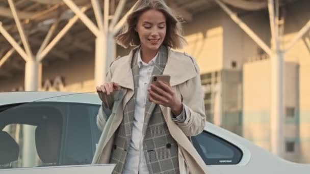 空港駐車場の車の近くに屋外に立っている間 スマートフォンを使用して離れて見てコートで陽気なブロンドのビジネス女性 — ストック動画