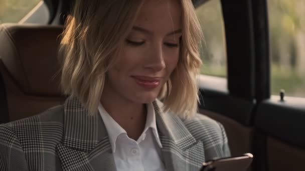 車の中で座っている間にスマートフォンを使用してコートで陽気なブロンドのビジネス女性のクローズアップビュー — ストック動画