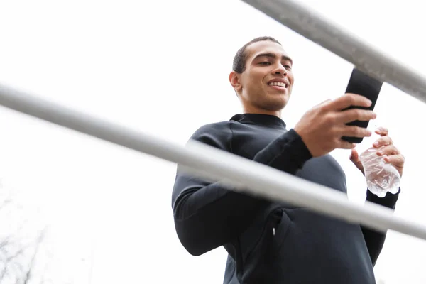 Foto de deportista sonriente guapo usando teléfono celular y sosteniendo la botella — Foto de Stock