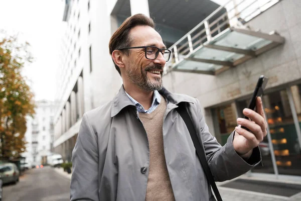 Εικόνα του χαρούμενου επιχειρηματία που χρησιμοποιεί κινητό τηλέφωνο και χαμογελά ενώ στέκεται — Φωτογραφία Αρχείου