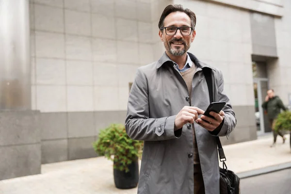 Εικόνα χαρούμενου επιχειρηματία που χρησιμοποιεί κινητό τηλέφωνο και χαμογελά ενώ στέκεται — Φωτογραφία Αρχείου