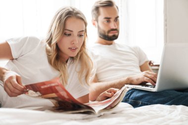 Couple indoors at home reading magazine using laptop computer.