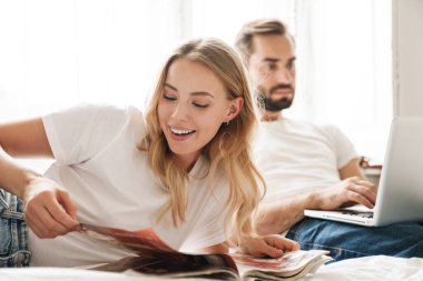 Couple indoors at home reading magazine using laptop computer.