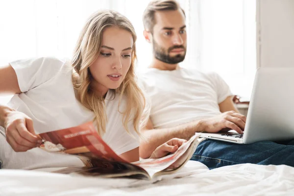 Couple indoors at home reading magazine using laptop computer. — 图库照片