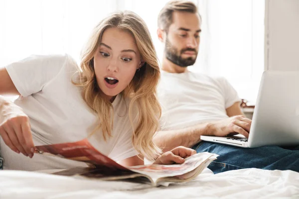 Couple indoors at home reading magazine using laptop computer. — 图库照片