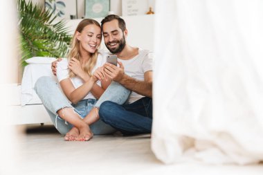 Loving couple sit on floor indoors using mobile phone.