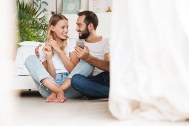 Loving couple sit on floor indoors using mobile phone.