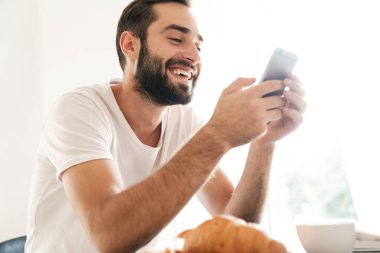 Man indoors at home have a breakfast using mobile phone.
