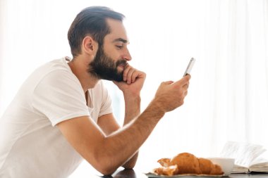 Man indoors at home have a breakfast using mobile phone.