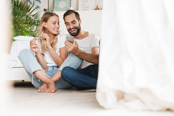 Loving couple sit on floor indoors using mobile phone. — Stock fotografie