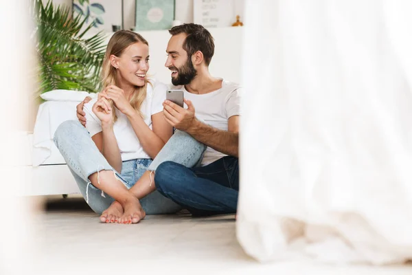 Loving couple sit on floor indoors using mobile phone. — Stock fotografie