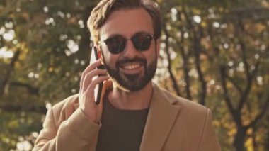 Palto ve güneş gözlüklü mutlu sakallı adam akıllı telefonla konuşuyor ve dışarıdaki parktaki kameraya bakıyor.