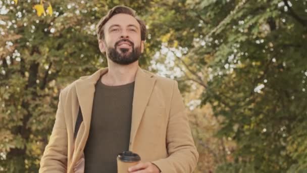 エレガントな髭を生やした男がコートを着て公園でコーヒーを飲みながら歩く屋外 — ストック動画