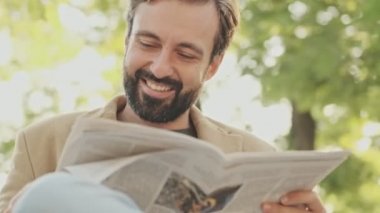 Gülümseyen, ceketli zarif sakallı adam elinde gazete tutarken ve parkta otururken başka tarafa bakıyor.