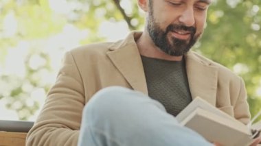 Paltolu zarif sakallı adam parkta otururken kitap okuyor.