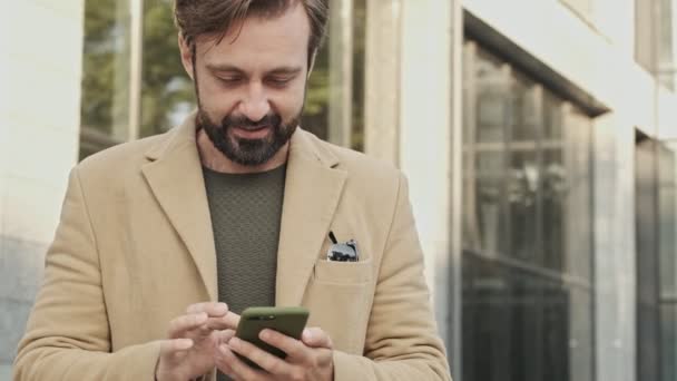 Uśmiechnięty Elegancki Brodaty Mężczyzna Płaszczu Pomocą Smartfona Zewnątrz — Wideo stockowe