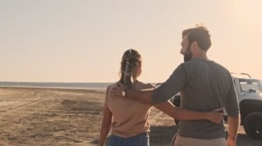 Mutlu, gülümseyen genç bir çiftin arka manzarası. Yazın göl kenarında birbirlerine sarılırlarken kadın ve erkek yürüyorlar.