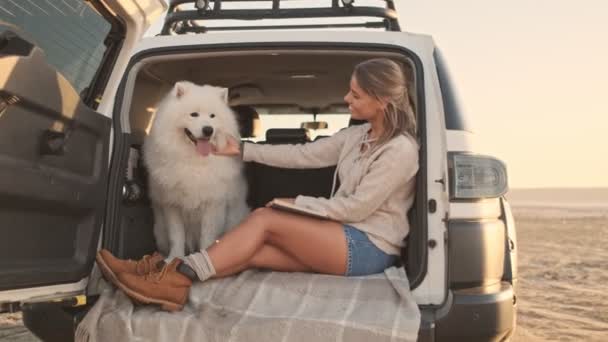 A nice young woman is petting a dog while reading a book in the trunk outdoors in summer. The camera moves away — Stock Footage Nice Young Woman Petting Dog While Reading Book Trunk Outdoors Royalty Free Stock Footage