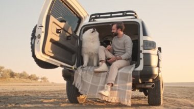 Yakışıklı bir genç adam yazın açık havada akıllı telefonunu kullanırken bagajında bir köpekle oturuyor.
