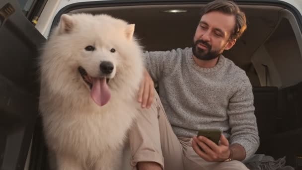 夏に屋外でスマートフォンを使用している間ハンサムな若い男がトランクの中に大きな白い犬をペッティングしています — ストック動画