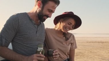 Olumlu gülümseyen genç bir çift, yaz mevsiminde gölde arazi aracının yanında dururken çay ya da kahve içiyorlar.