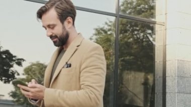 Palto giymiş, akıllı telefon kullanan ve dışarıda yürürken yüzünü çeviren zarif sakallı adamın yan görüntüsü.