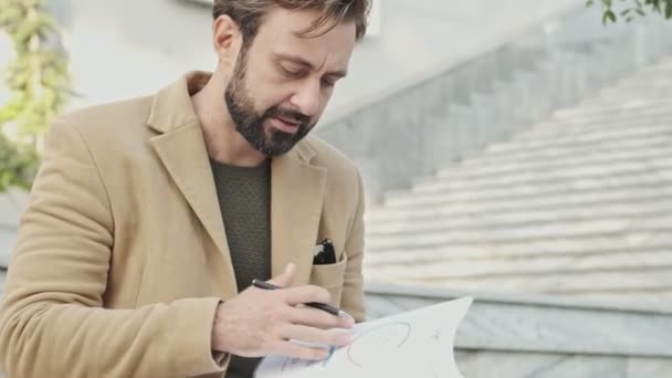 外の階段の近くに座っている間 コートの読書文書でエレガントな髭の男を集中 — ストック動画